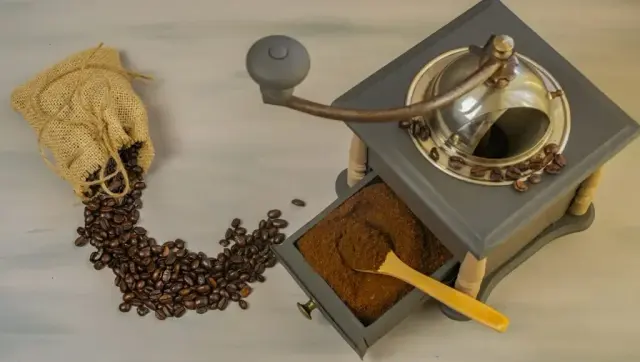 Roasted coffee beans spilling from a burlap sack beside a wooden hand grinder filled with ground coffee